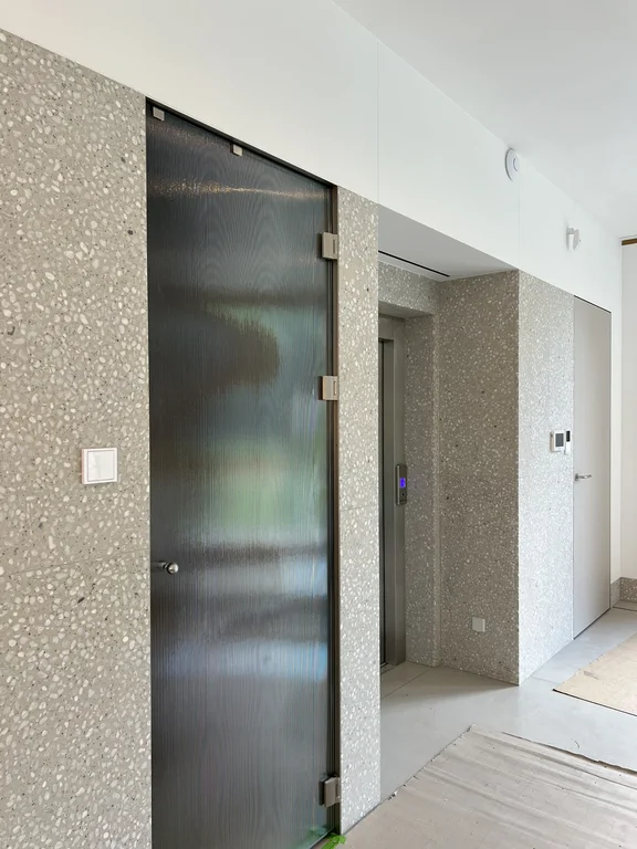 Szklane drzwi, winda i jasne ściany wykończone lastrykiem w nowoczesnym holu, widoczna winda 