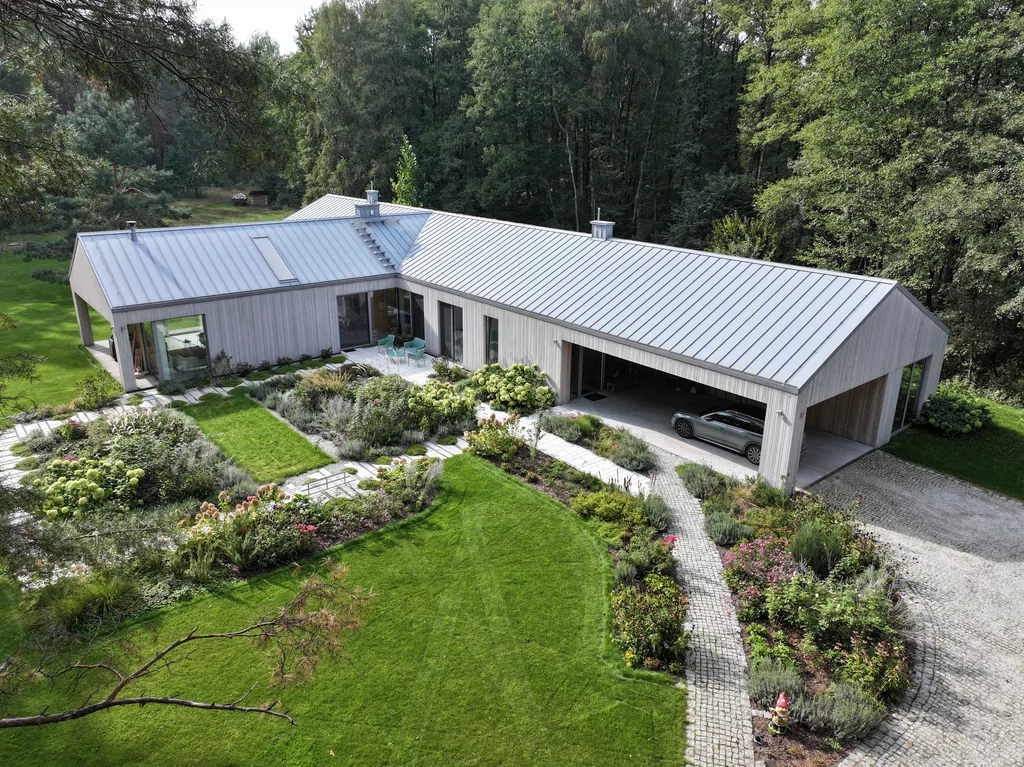 Szeroki kadr z drona: dom parterowy z drewnianą elewacją i dachem na rąbek, reprezentacyjny ogród z rabatami i krętą ścieżką prowadzącą do podcienia/wiaty.