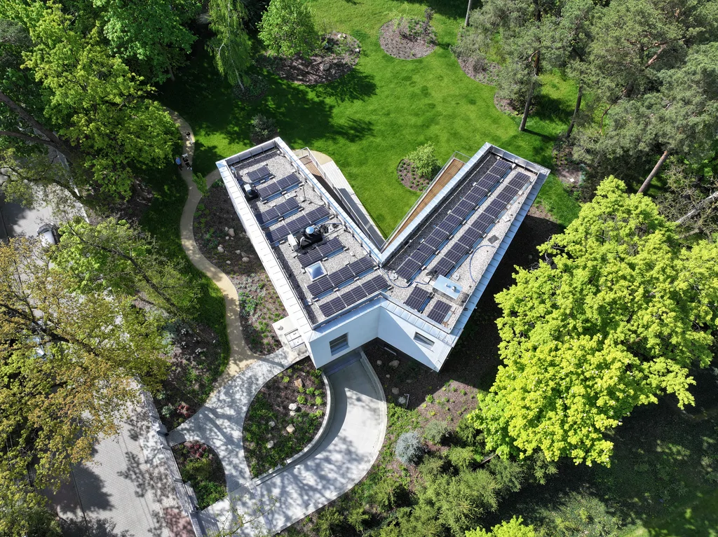 Pionowy widok z drona na bryłę domu w kształcie litery V z panelami fotowoltaicznymi, zakrzywionym podjazdem i ścieżkami wśród zieleni