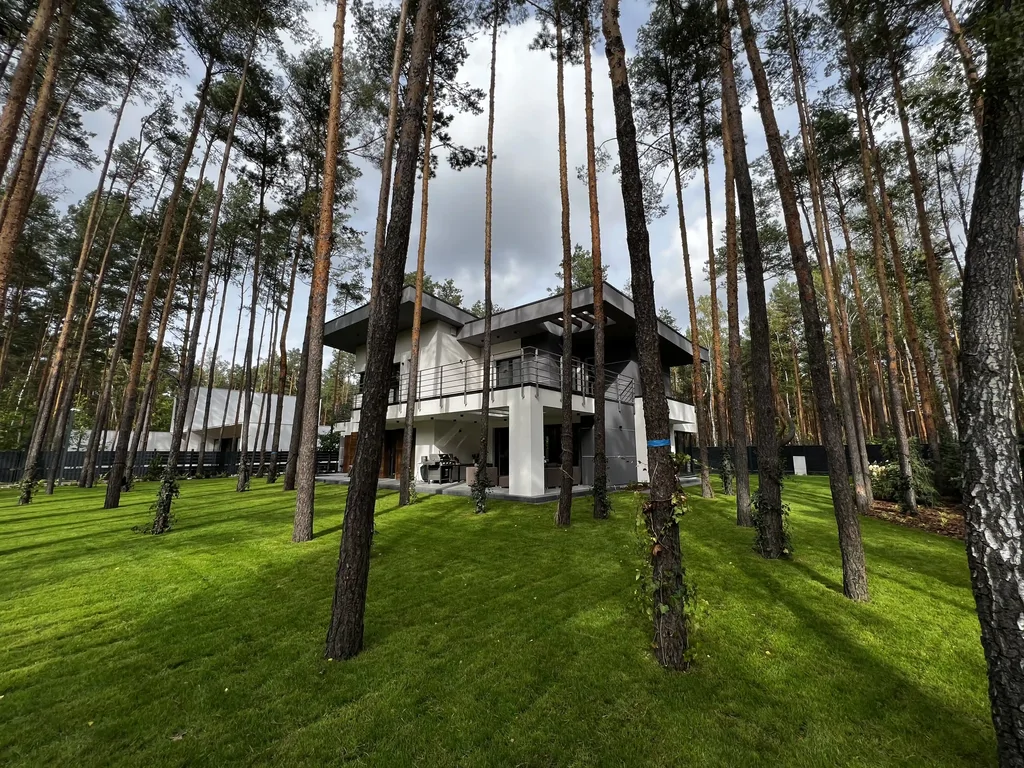 Dom widziany przez sosny: zielony trawnik, duże okapy, taras i balkon dookoła budynku