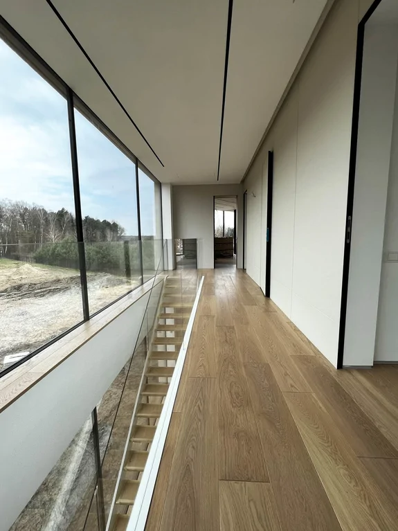 Korytarz na piętrze z balustradą szklaną i widokiem na otwartą klatkę schodową. Duże przeszklenie doświetla strefę komunikacji.