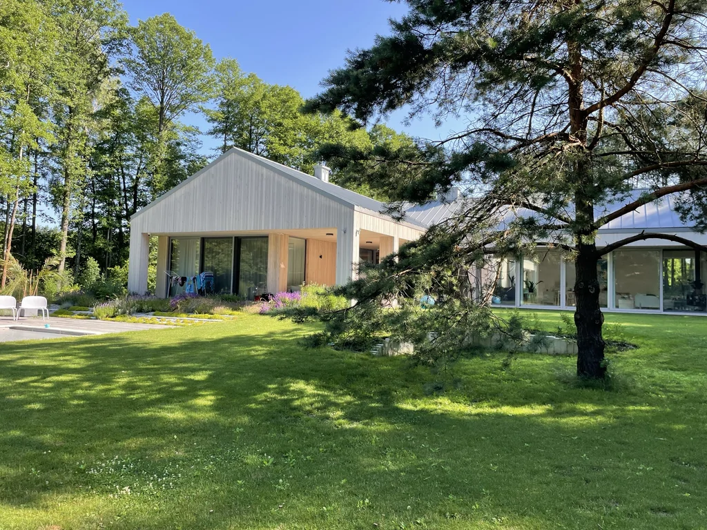 Ujęcie ogrodu z dużym drzewem na pierwszym planie: parterowy dom z drewnianą elewacją, dachem na rąbek i dużymi przeszkleniami, otoczenie leśne.