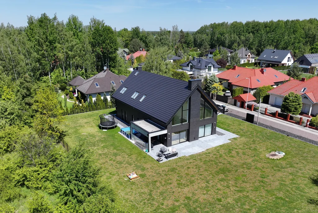 Ujęcie z drona pod kątem: dom w Ruścu z ciemnej cegły, taras i otoczenie działki z zabudową sąsiednią.