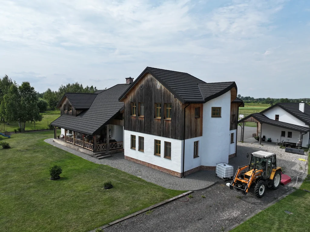 Widok z drona na boczną elewację z długą, zadaszoną werandą; obok widoczny sprzęt gospodarczy