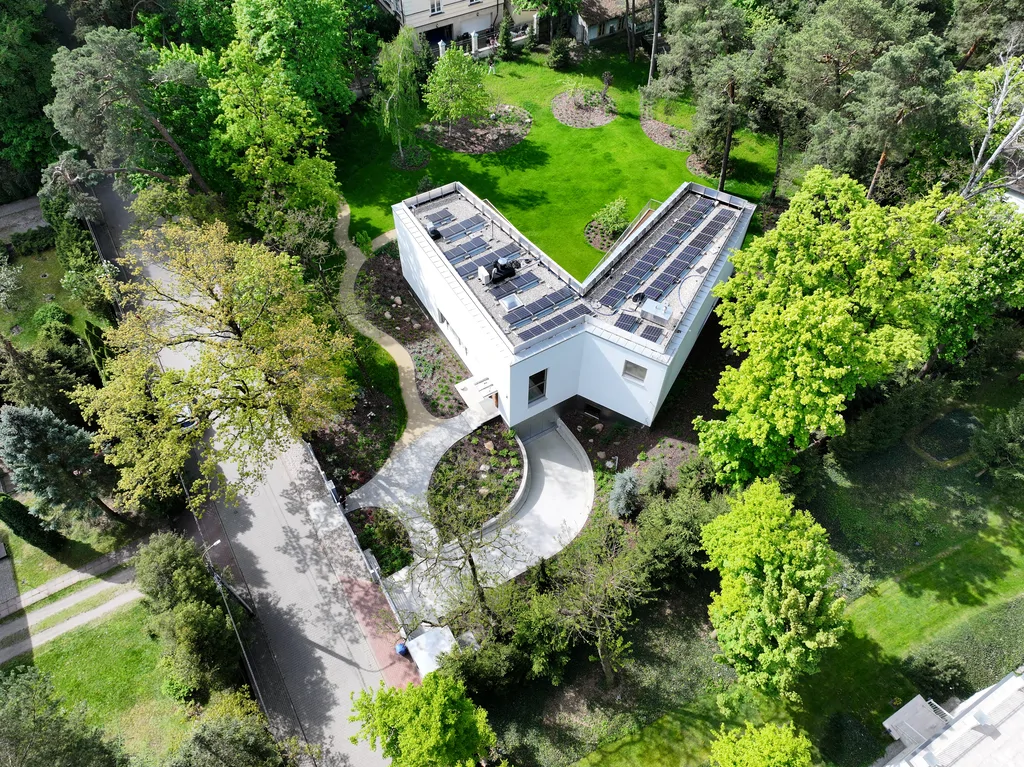 Szerokie ujęcie z lotu ptaka na frontową część posesji z nowoczesnym białym domem, półkolistym podjazdem i dużym ogrodem otoczonym drzewami