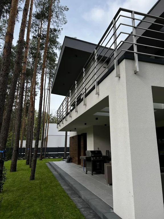 Perspektywa tarasu wzdłuż elewacji: grill, podcień i balkon powyżej, sosny przy ogrodzeniu