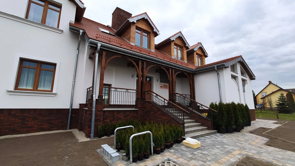 Frontowe schody i wejście do domu zakonnego w Ostródzie pod drewnianym gankiem, z donicami z kwiatami