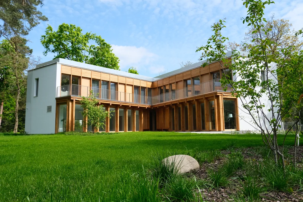 Widok od strony ogrodu na nowoczesny dom w kształcie litery L z drewnianą elewacją, balkonem i rozległym trawnikiem