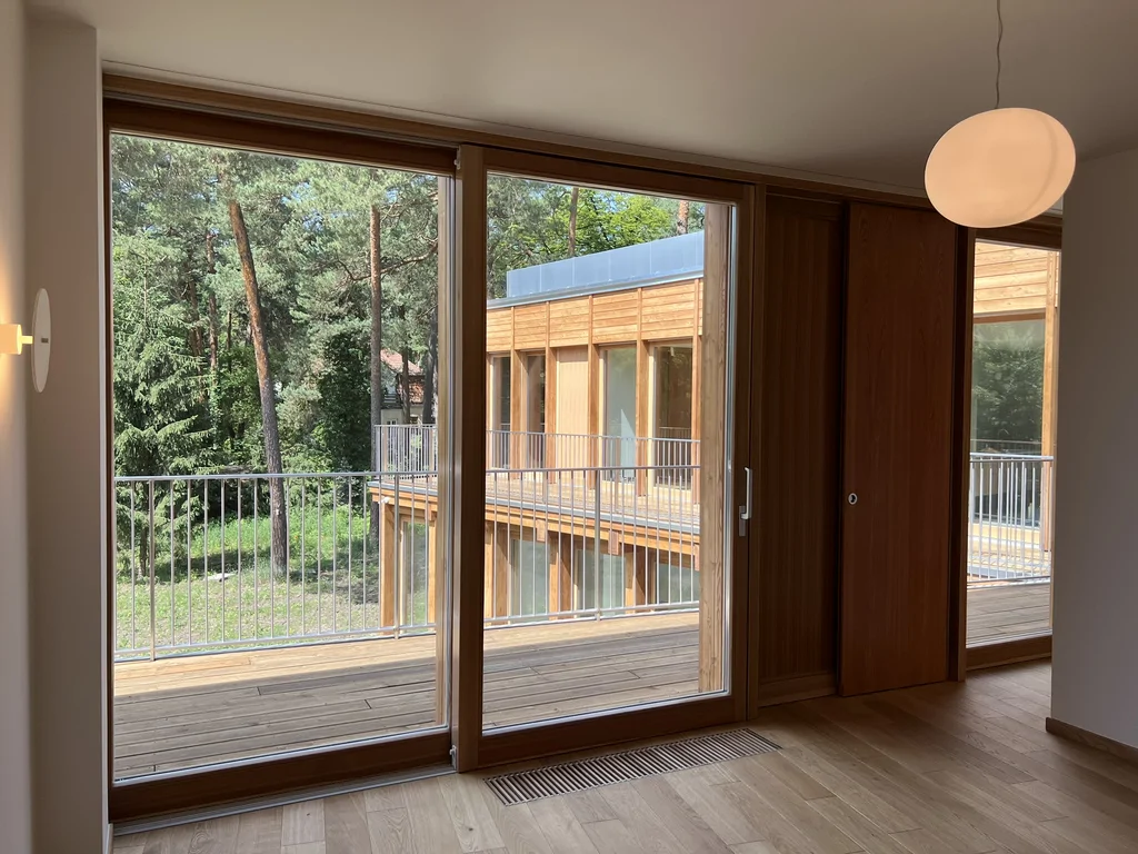Pokój z dużym przeszkleniem i widokiem na drewniany taras oraz sąsiednie skrzydło budynku