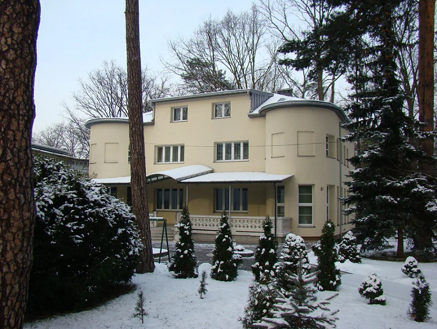 Widok frontu budynku w zimowej scenerii; zaokrąglone skrzydła, schody wejściowe i nasadzenia w ogrodzie