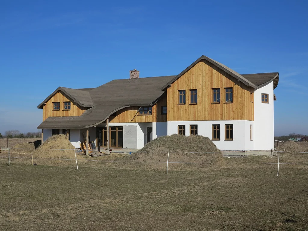 Boczny widok domu w trakcie budowy; drewniane szczyty, biały tynk i pryzmy ziemi na placu