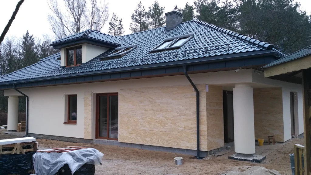 Jasna elewacja strefa wejścia, dach pokryty blachą na rąbek, okrągły słup, brązowa stolarka okien, komin obmurowany klinkierem