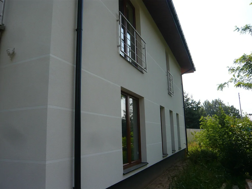 Boczna elewacja domu – jasny tynk z poziomymi pasami i metalowa balustrada przy oknie/wyjściu