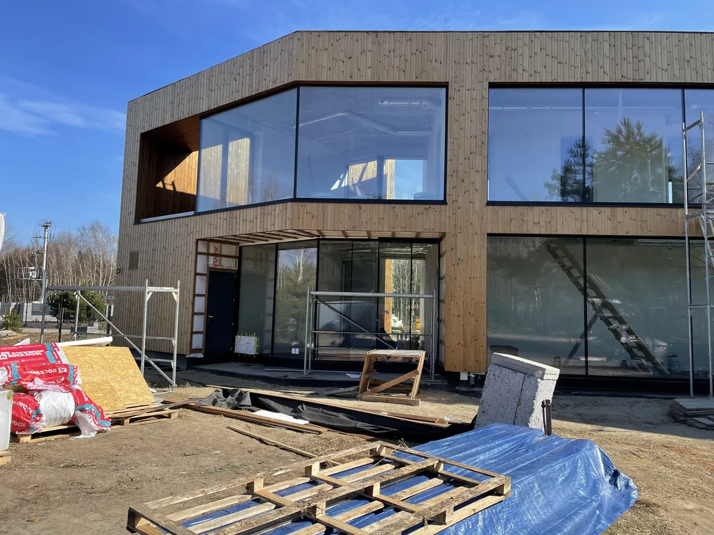 Elewacja drewniana z dużym narożnym przeszkleniem i strefą wejściową; budynek w zaawansowanym etapie wykończenia.