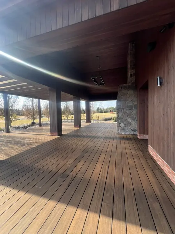 Zadaszona część tarasu po remoncie z drewnopodobną podłogą, kamiennym kominkiem i słupami konstrukcyjnymi.