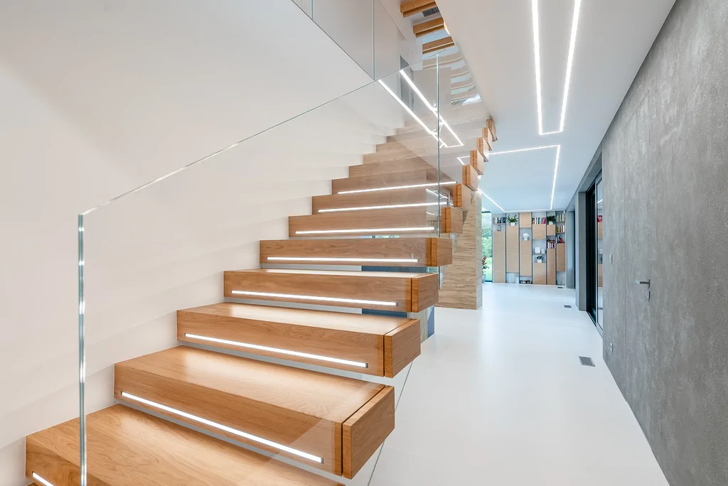 Schody dębowe ze szklaną balustradą i liniowym oświetleniem sufitu w strefie dziennej.