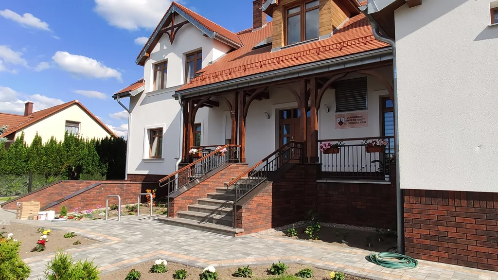 Tylna elewacja domu zakonnego w Ostródzie w słoneczny dzień, z nasadzeniami i utwardzonym podjazdem