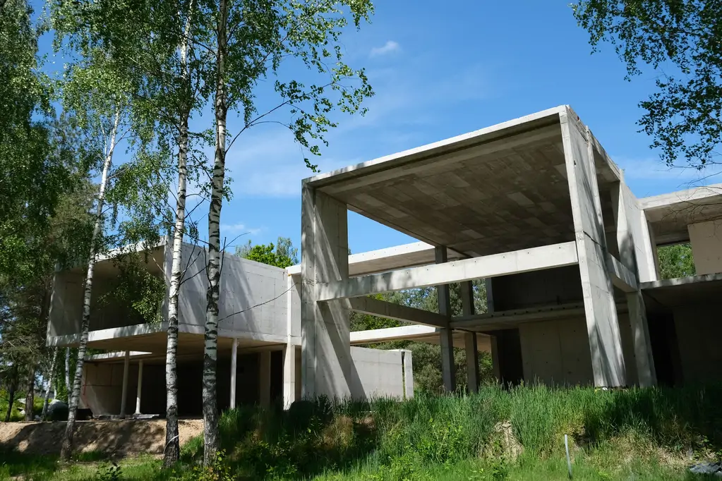 Widok na nowoczesny dom żelbetowy w trakcie budowy, otoczony brzozami i wysoką zielenią.