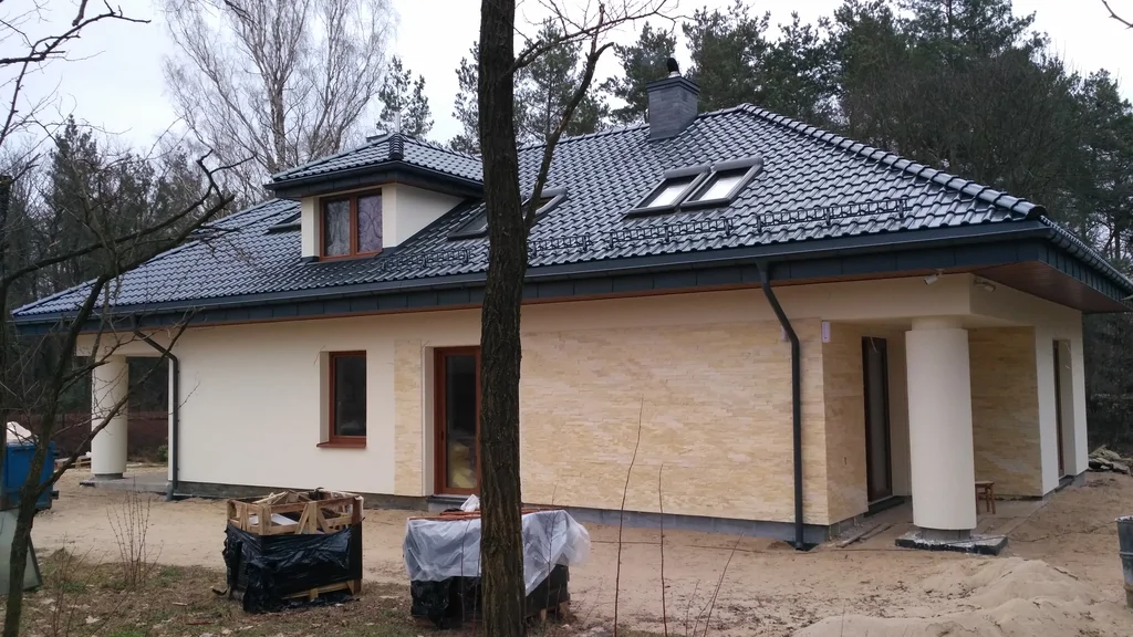 Jasna elewacja strefa wejścia, dach pokryty blachą na rąbek, okrągły słup, brązowa stolarka okien, komin obmurowany klinkierem