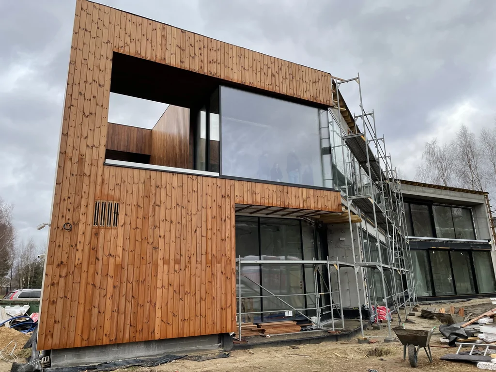 Narożna bryła w drewnie z dużym oknem i wyciętą wnęką; prace wykończeniowe i rusztowanie na sąsiedniej części budynku.