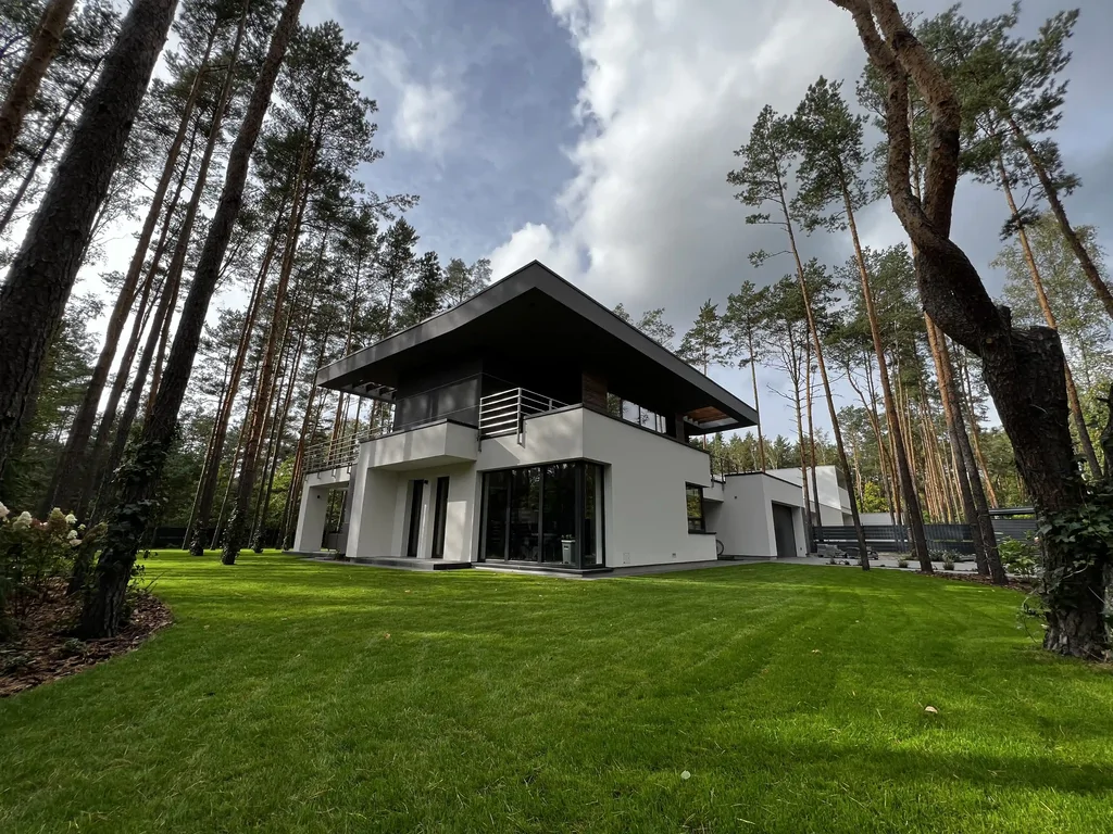 Szeroki kadr domu na leśnej działce: duży trawnik, biała i grafitowa elewacja, przeszklony narożnik