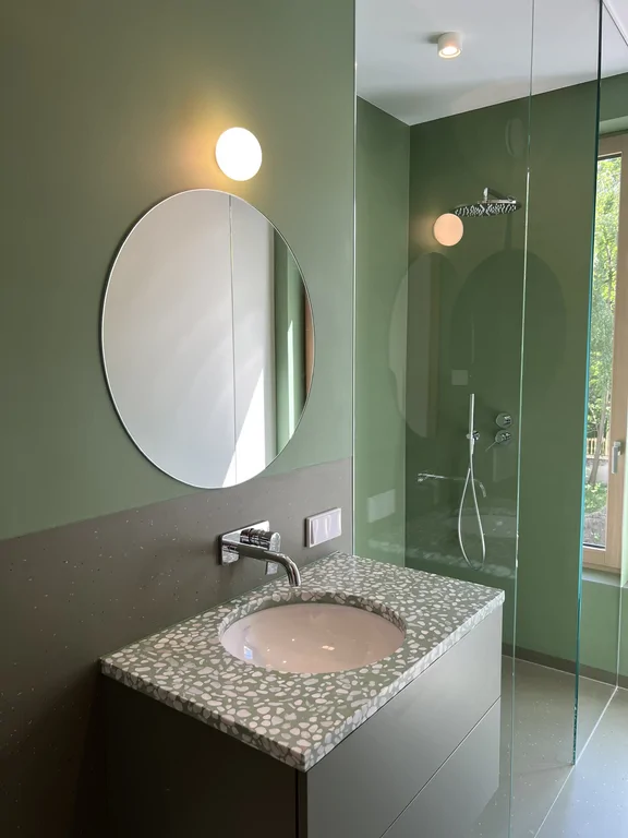 Zielona łazienka z umywalką, okrągłym lustrem i drewnianymi drzwiami