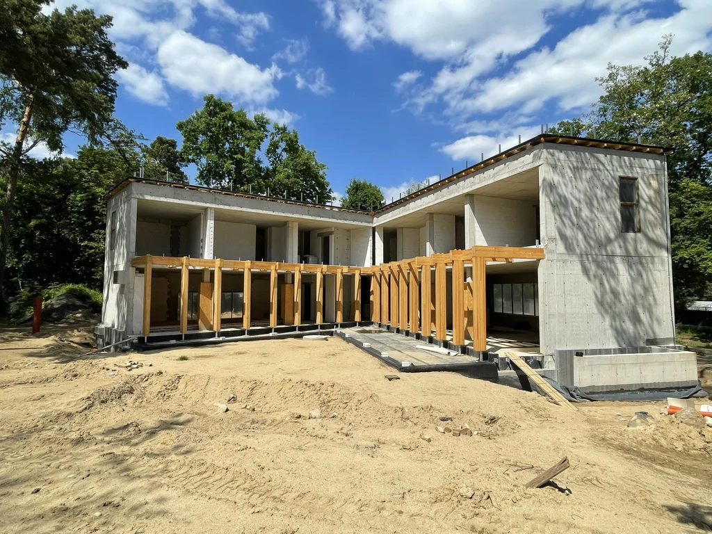 Dom jednorodzinny w stanie surowym z żelbetową konstrukcją, dużymi przeszkleniami i drewnianą pergolą od strony ogrodu