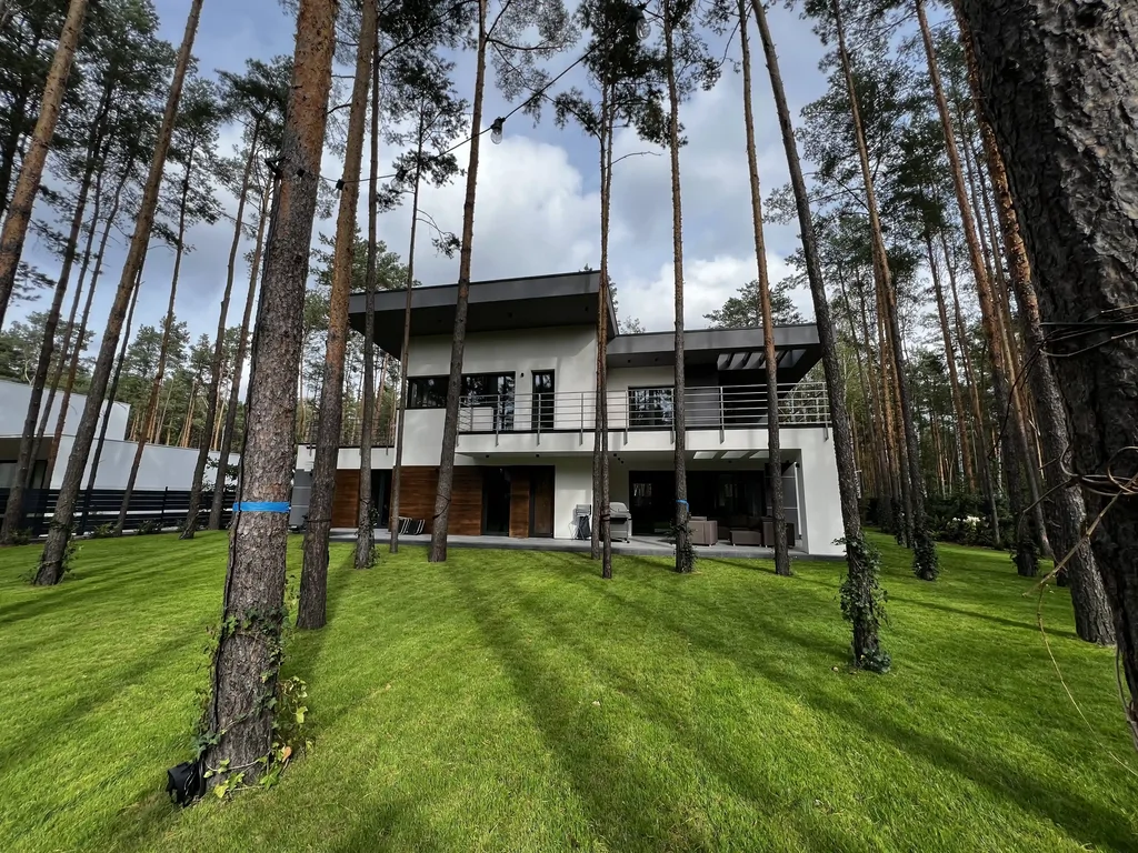 Elewacja od ogrodu wśród wysokich sosen: duży trawnik, balkon na całej długości i taras pod zadaszeniem