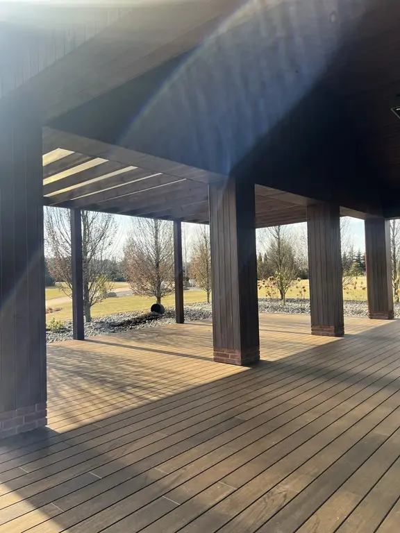 Zadaszony taras po remoncie z drewnopodobną posadzką, ciemnymi słupami i widokiem na ogród w słońcu.