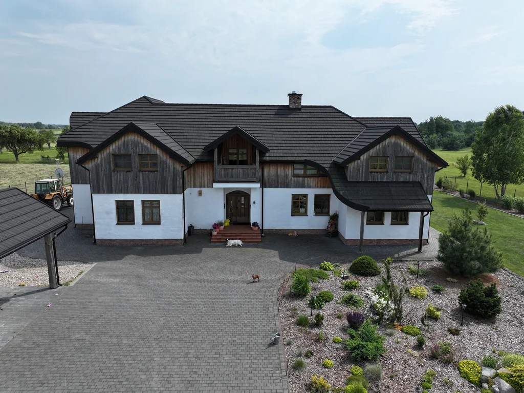 Ujęcie z drona na frontową elewację domu z ogrodem żwirowym i niskimi krzewami oraz podjazdem z kostki