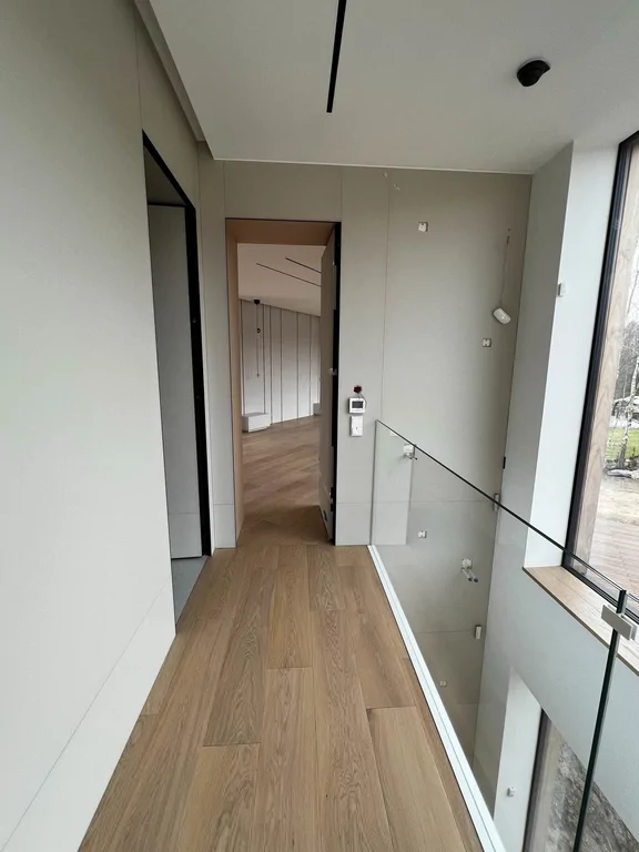 Korytarz na piętrze z dębową podłogą i szklaną balustradą nad dwukondygnacyjną przestrzenią.