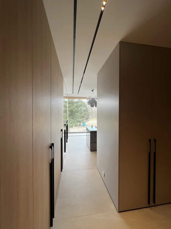 Korytarz “w ramie” wysokich szaf, prowadzący w stronę strefy dziennej. Oświetlenie liniowe porządkuje sufit.