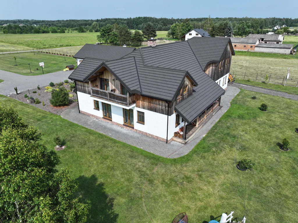 Widok z drona na boczną elewację domu z balkonem i tarasem oraz rabaty roślinne przy podjeździe