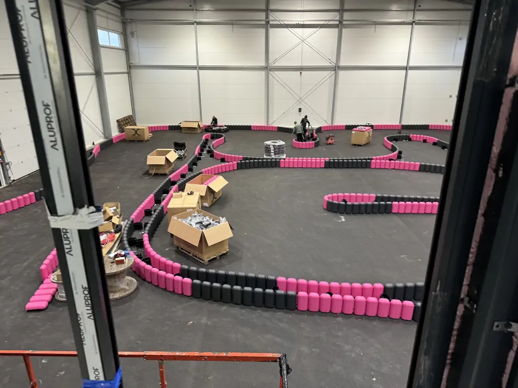 Widok z antresoli na halę z torem gokartowym w trakcie montażu, z różowo-czarnymi bandami i pracującą ekipą.