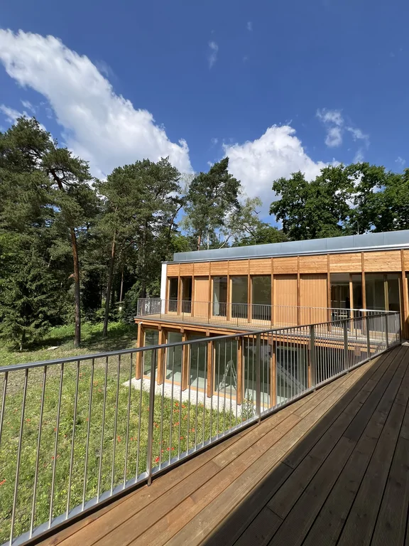 Widok z drewnianego tarasu na drugie skrzydło nowoczesnego domu i otaczające drzewa