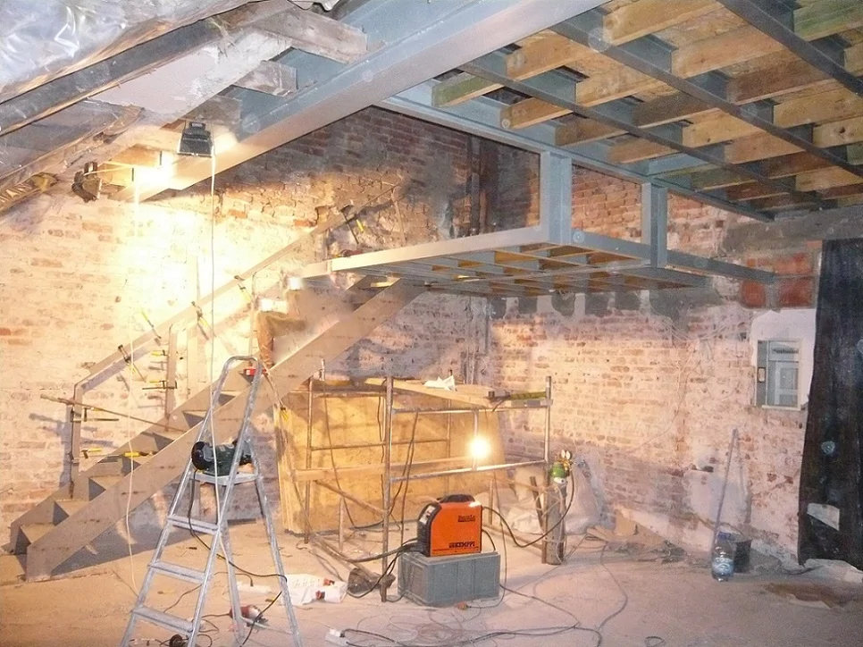 Prace montażowe przy stalowej antresoli i schodach, oświetlenie robocze i spawarka w surowym wnętrzu