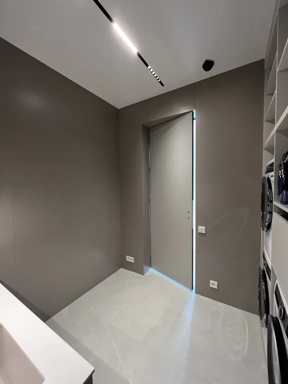 Minimalistyczna pralnia w stonowanych szarościach, z jasną posadzką i liniowym oświetleniem sufitowym.