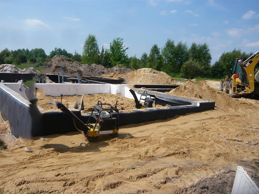 Zasypka i zagęszczanie gruntu przy fundamentach; widoczna izolacja i prace ziemne koparką