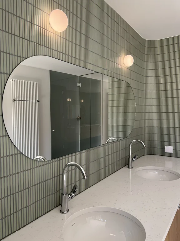 Detal łazienki z podwójną umywalką, dużym lustrem i zieloną ścianą z pionowych płytek