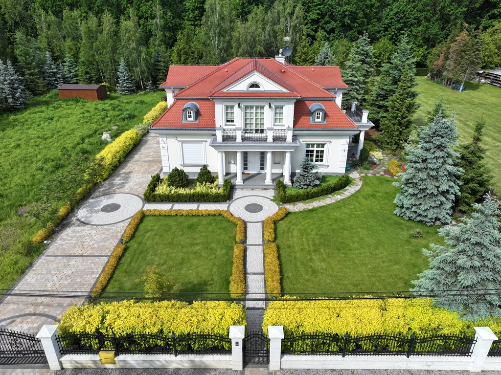 Widok z drona na dom jednorodzinny w stylu dworkowym z białą elewacją z boniami, kolumny okrągłe w strefie wejścia, na balkonie również kolumny i stalowa barierka, pokrycie dachu karpiówka, na dachu widoczne lukarny, przy domu kostka, działka otoczona drzewami 