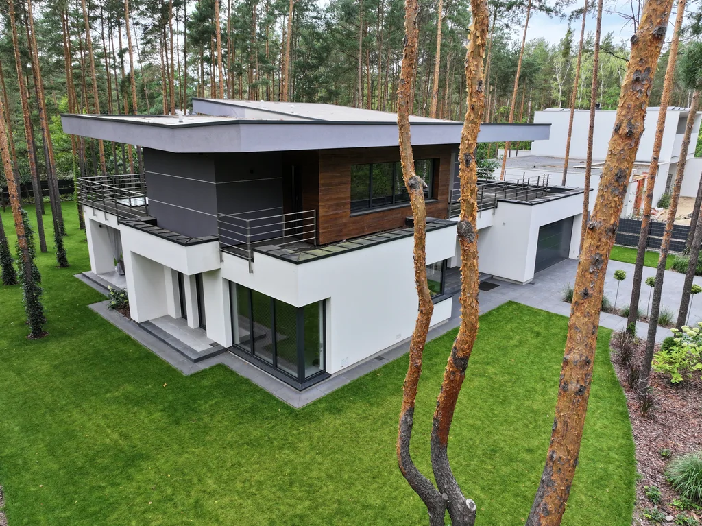 Ujęcie z drona z boku: biała elewacja, narożne przeszklenia i tarasy okalające budynek na tle lasu