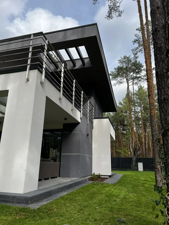 Zbliżenie na balkon i okap z pergolą; grafitowo-biała elewacja i leśne otoczenie