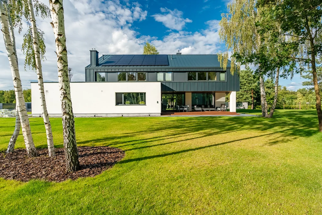 Tylna elewacja nowoczesnego domu z dużym ogrodem, tarasem i przeszkloną strefą dzienną.