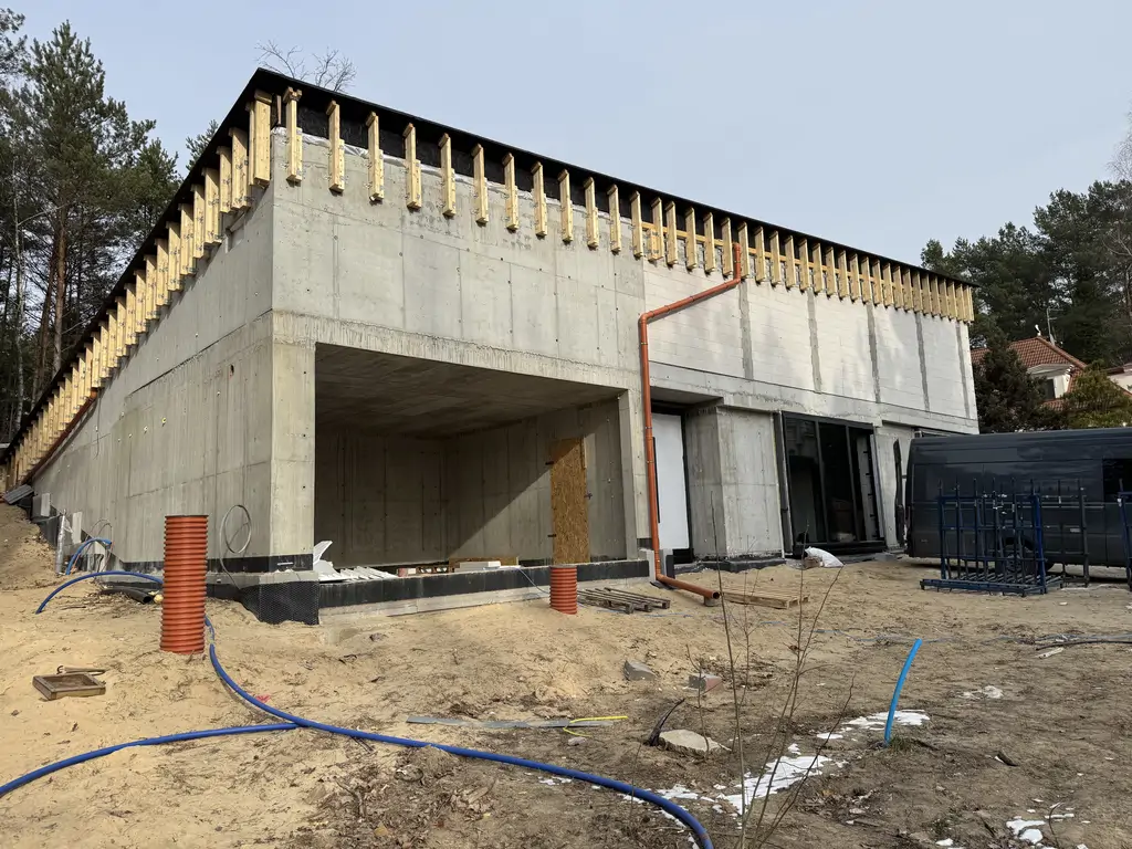 Nowoczesny budynek w stanie surowym z żelbetową konstrukcją, otwartym podcieniem i pochyłym stropodachem.