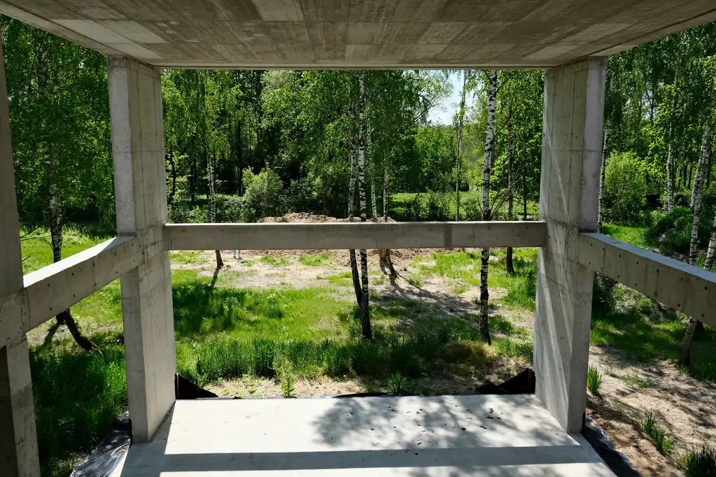 Widok przez otwartą żelbetową konstrukcję domu na zieloną działkę i drzewa w tle.