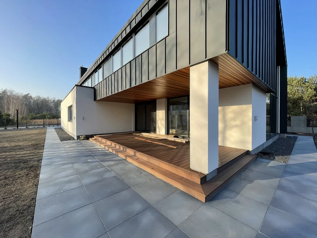 Taras nowoczesnego domu pod nadwieszoną bryłą, drewniana podłoga i duże przeszklenia