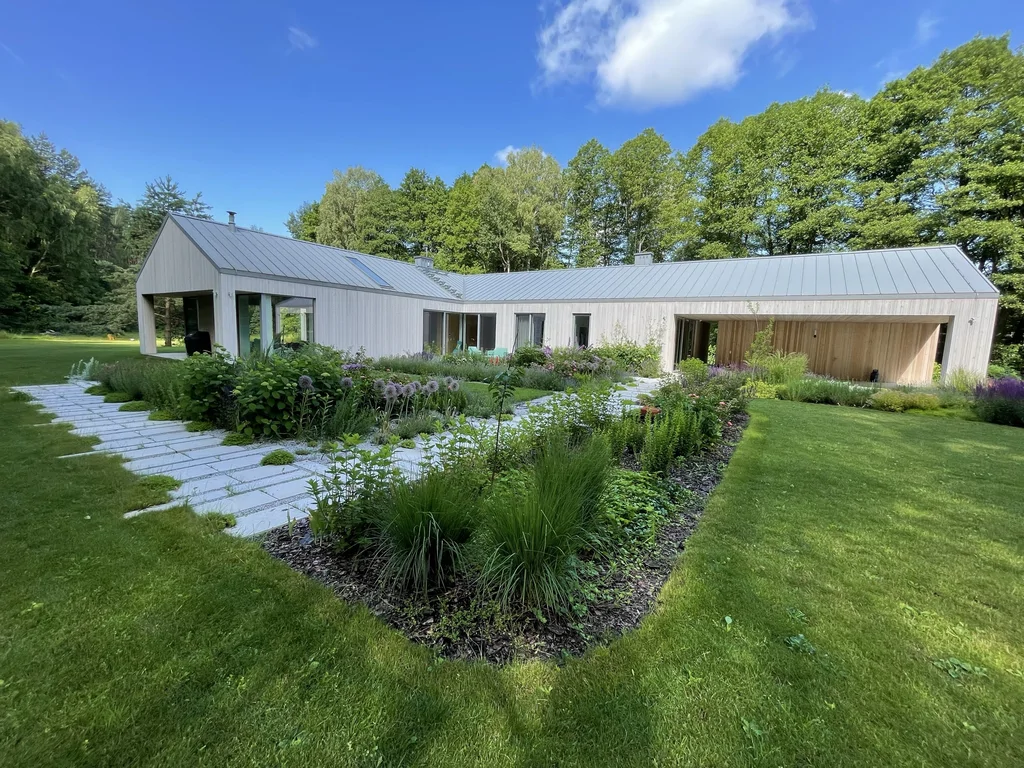 Widok ogólny domu parterowego: jasna elewacja drewniana, dach na rąbek, duże przeszklenia i podcień, a na pierwszym planie ogród z rabatami i ścieżką z płyt.