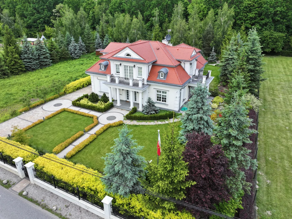 Ujęcie z drona przedstawiające dużą posesję z reprezentacyjnym domem, rozległym ogrodem, geometrycznymi ścieżkami i wysokimi drzewami wokół działki.
