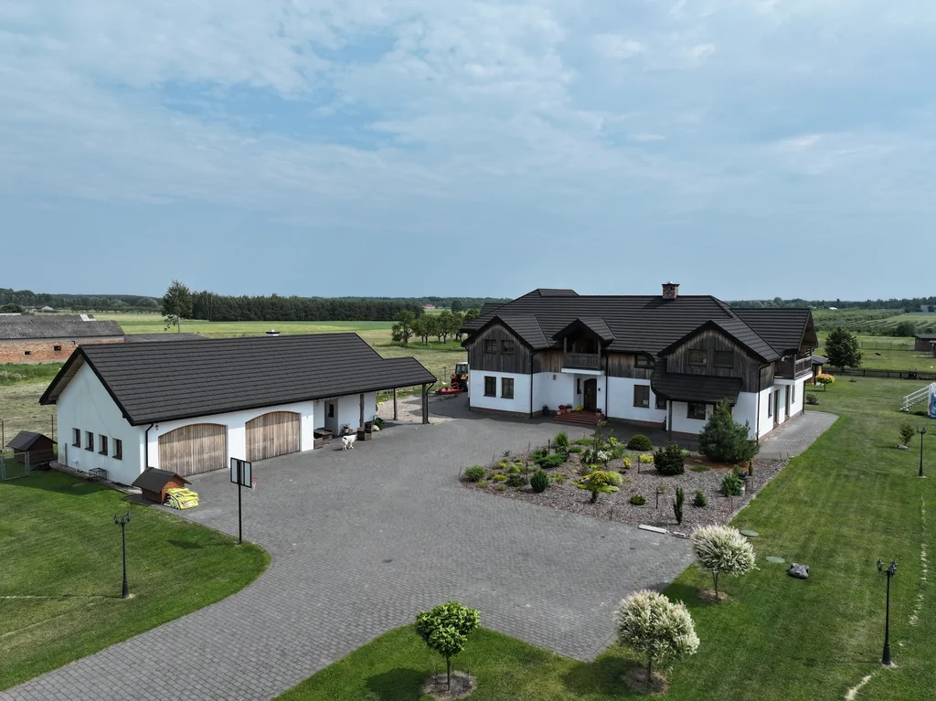 Widok z drona na dom z białą elewacją i drewnianą okładziną oraz budynek garażowy i szeroki podjazd z kostki