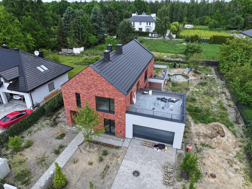 Ujęcie z drona na front domu z ceglaną elewacją, dużym tarasem dachowym nad garażem i podjazdem